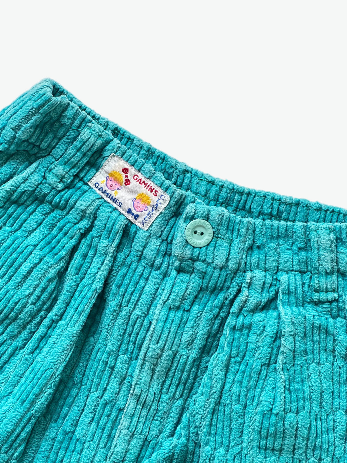 Close-up of teal corduroy trousers with a brand label on a white background