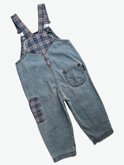 Kids vintage blue denim dungarees with check details and badge on chest pocket laid flat on a white background.