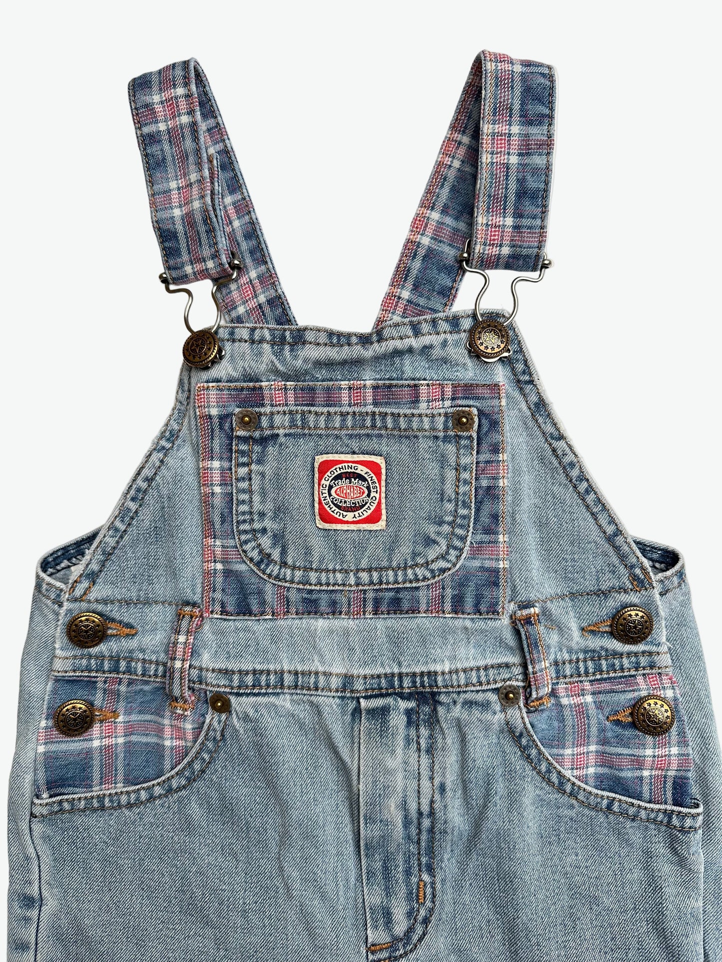 Kids vintage blue denim dungarees with check details and badge on chest pocket laid flat on a white background.