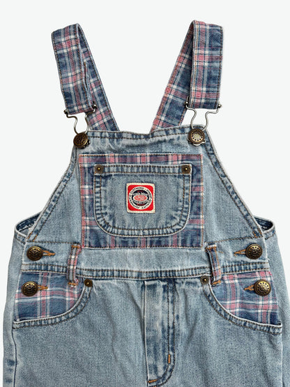 Kids vintage blue denim dungarees with check details and badge on chest pocket laid flat on a white background.