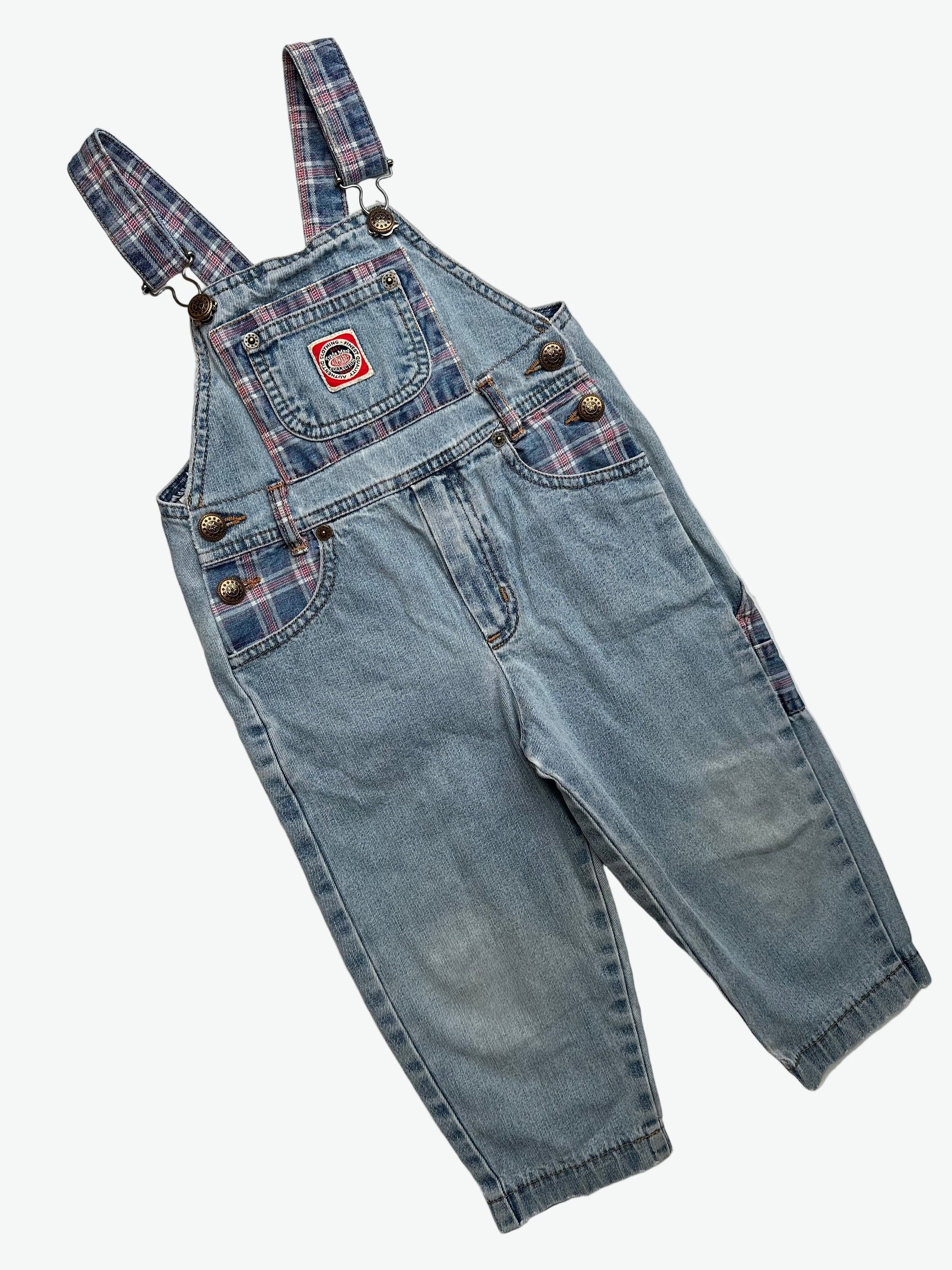 Kids vintage blue denim dungarees with check details and badge on chest pocket laid flat on a white background.