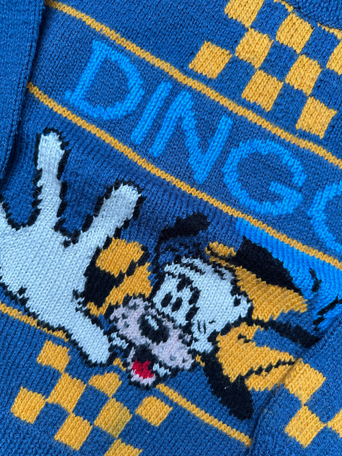  Close-up of a knit jumper with a dog illustration and 'Dingo' text on a blue and yellow checkered background.