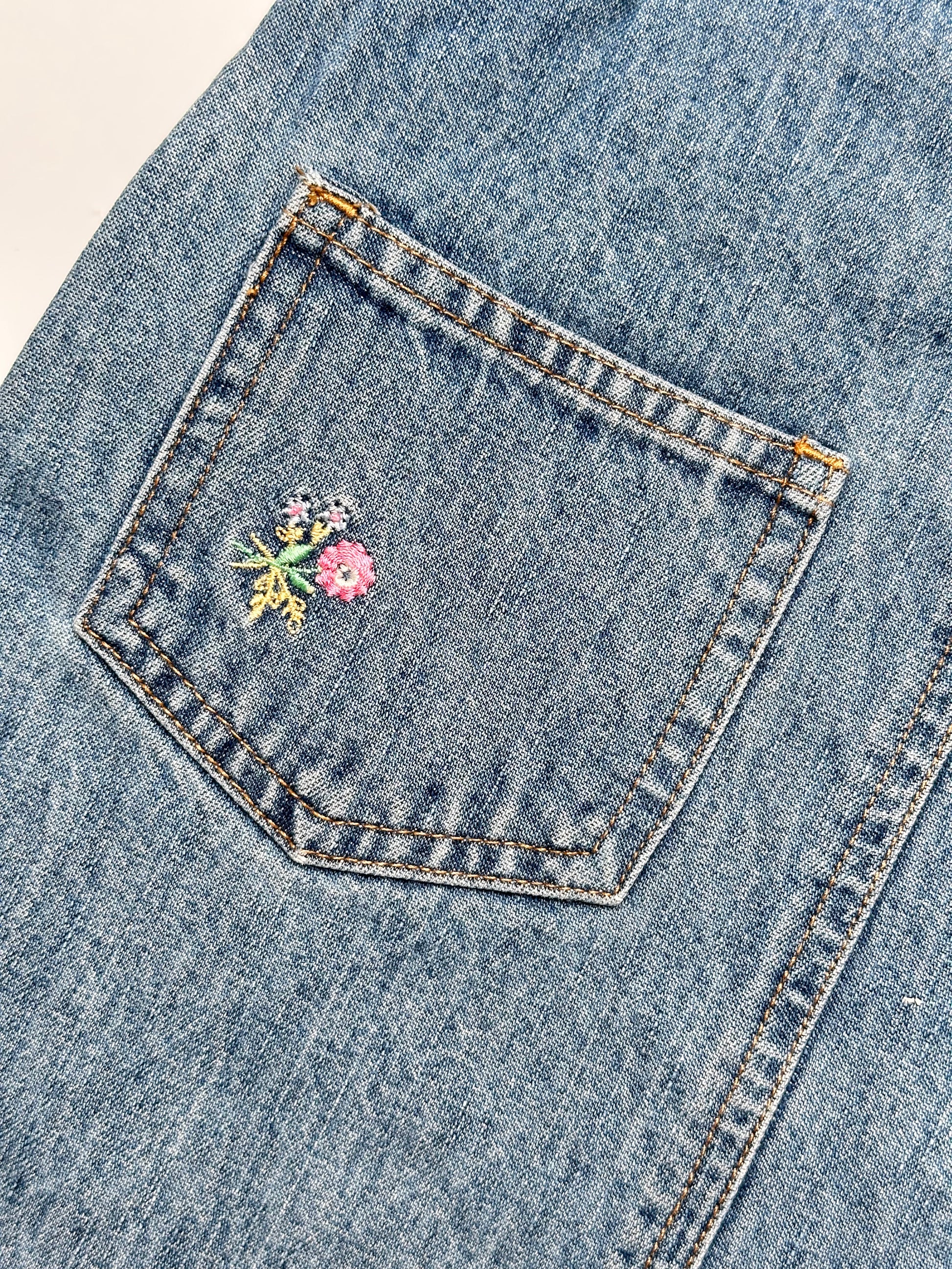Kids vintage OshKosh dungaree dress, featuring ditsy floral lining inside the top, delicate embroidered flowers, and playful patch details. Mid-wash denim with classic OshKosh branding, it’s finished with antique brass trimmings and sturdy dungaree buckles. Age 4 years.
