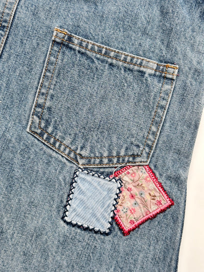 Kids vintage OshKosh dungaree dress, featuring ditsy floral lining inside the top, delicate embroidered flowers, and playful patch details. Mid-wash denim with classic OshKosh branding, it’s finished with antique brass trimmings and sturdy dungaree buckles. Age 4 years.