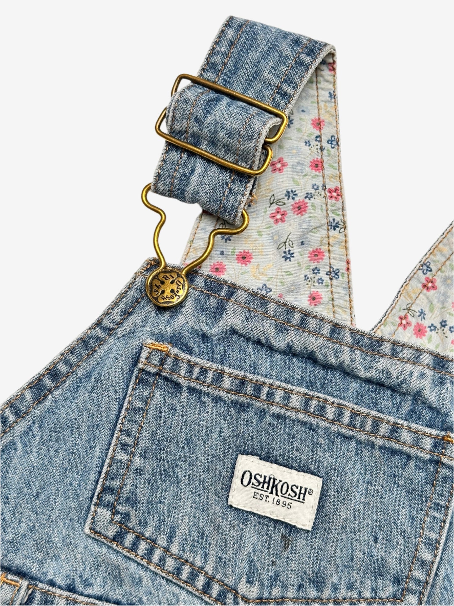 Kids vintage OshKosh dungaree dress, featuring ditsy floral lining inside the top, delicate embroidered flowers, and playful patch details. Mid-wash denim with classic OshKosh branding, it’s finished with antique brass trimmings and sturdy dungaree buckles. Age 4 years.