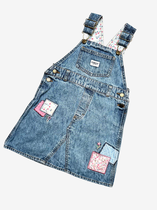 Kids vintage OshKosh dungaree dress, featuring ditsy floral lining inside the top, delicate embroidered flowers, and playful patch details. Mid-wash denim with classic OshKosh branding, it’s finished with antique brass trimmings and sturdy dungaree buckles. Age 4 years.